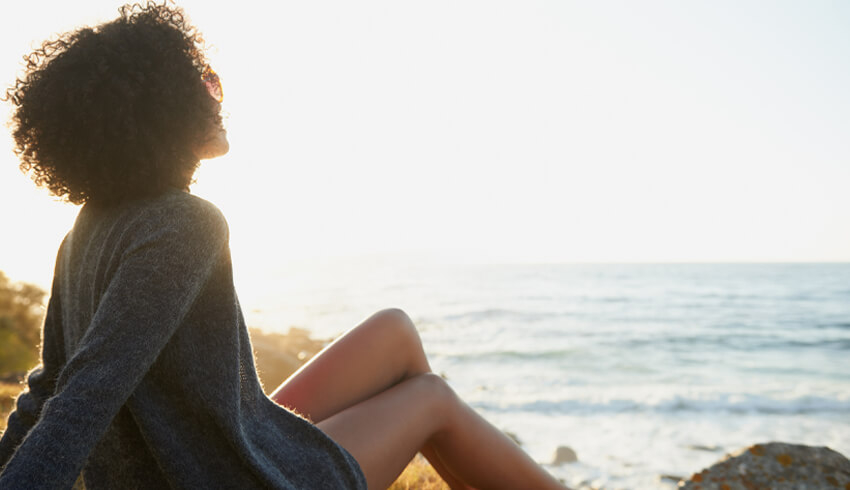 A woman sits on a hill overlooking waves rolling in on a beach on a sunny day